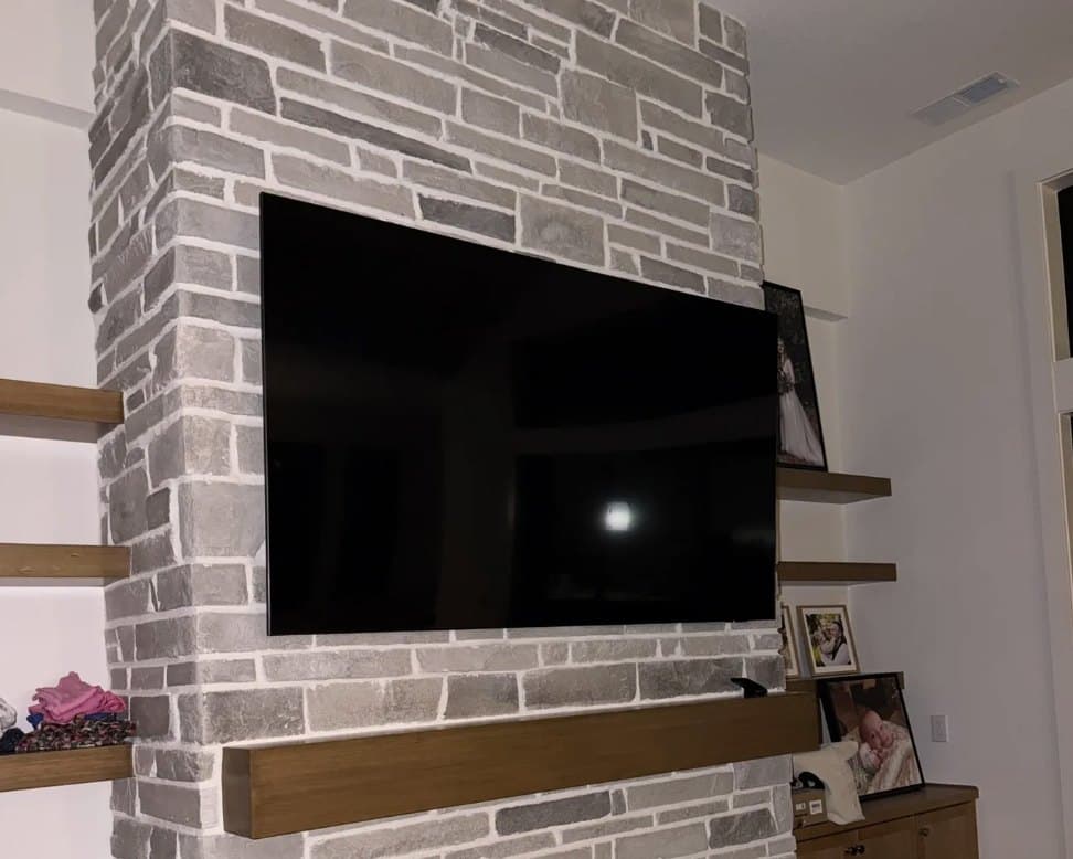Large TV mounted on a grey brick chimney breast above a solid wood mantel, sitting flush with no visible cables