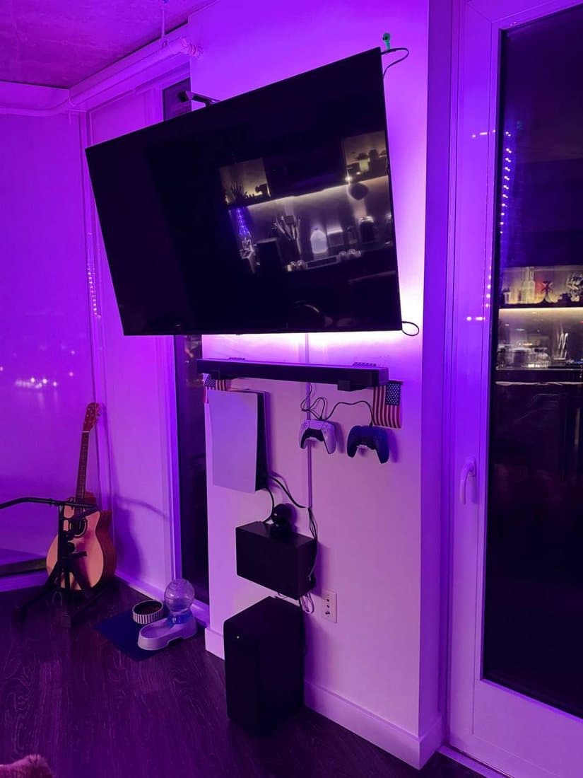 TV on a full-motion mount angled out from a corner, purple bias lighting behind the screen and a floating shelf holding game consoles and controllers